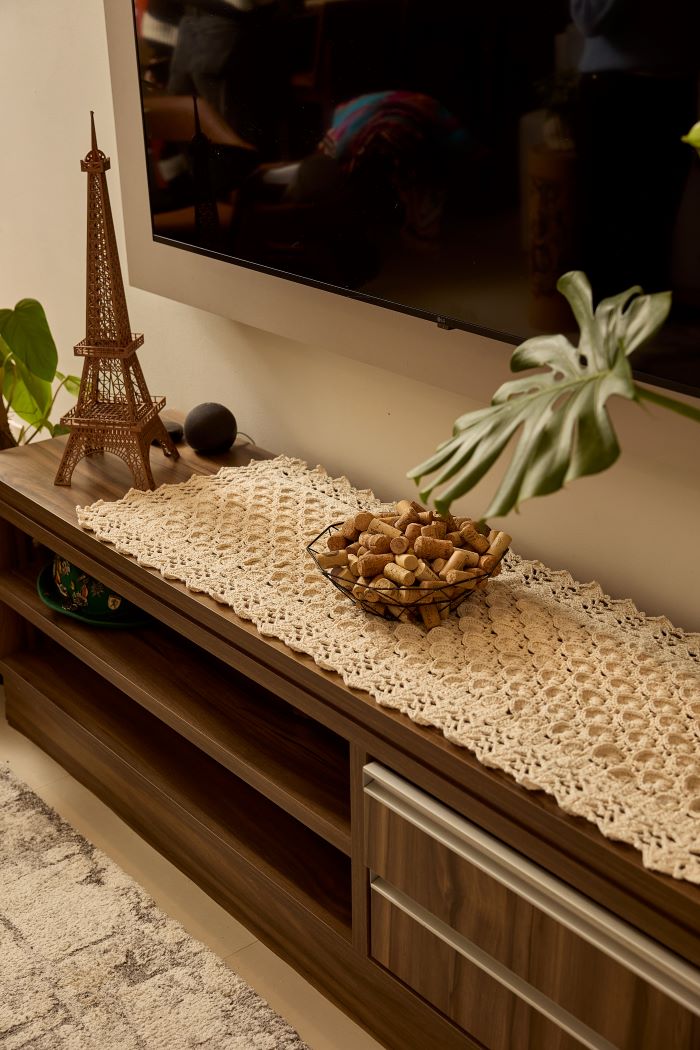 Circulo Barroco Natural Brilho - Natural Cotton Yarn Gold/Silver, 657m / 433m, 400g - A cream-colored crocheted lace runner tablecloth draped over a dark wood TV stand in a living room setting, styled with a small Eiffel Tower figurine, a bowl of decorative stones, and a potted monstera plant.