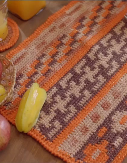 Círculo Barroco Maxcolor Sparkle – Cotton Yarn with Metallic Shine (200g) - A crocheted table runner with a geometric pattern in warm orange, beige, and brown tones, displayed on a wooden table with fruit and juice glasses.